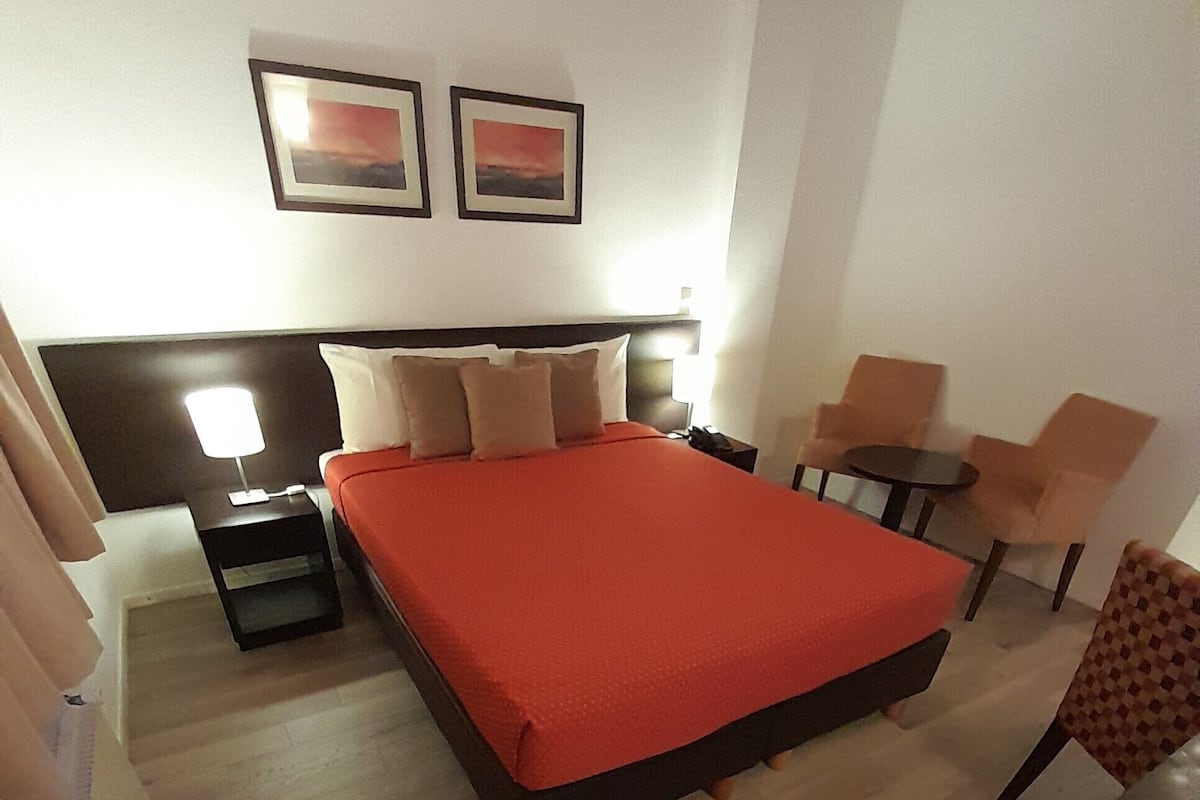 A hotel room with a large bed, a bedside table with a lamp, a small table with chairs, and two framed pictures on the wall.