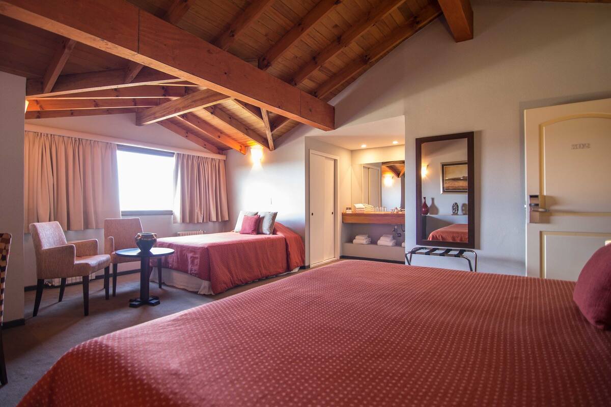 A hotel room with a wooden ceiling, two beds with red bedding, a chair, a small table, a mirror, and a window with curtains.