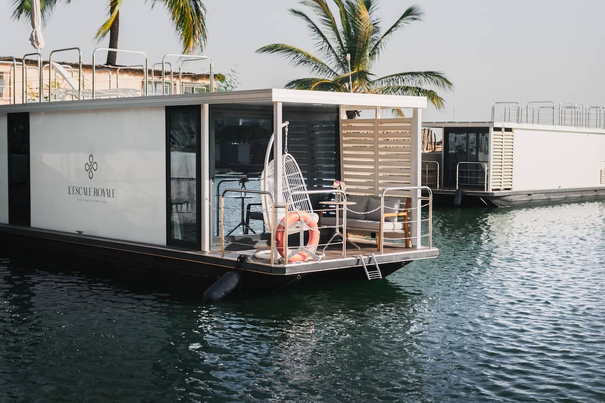 Uma embarcação flutuante com exterior branco, uma porta de vidro e área de estar com mesa e cadeiras.