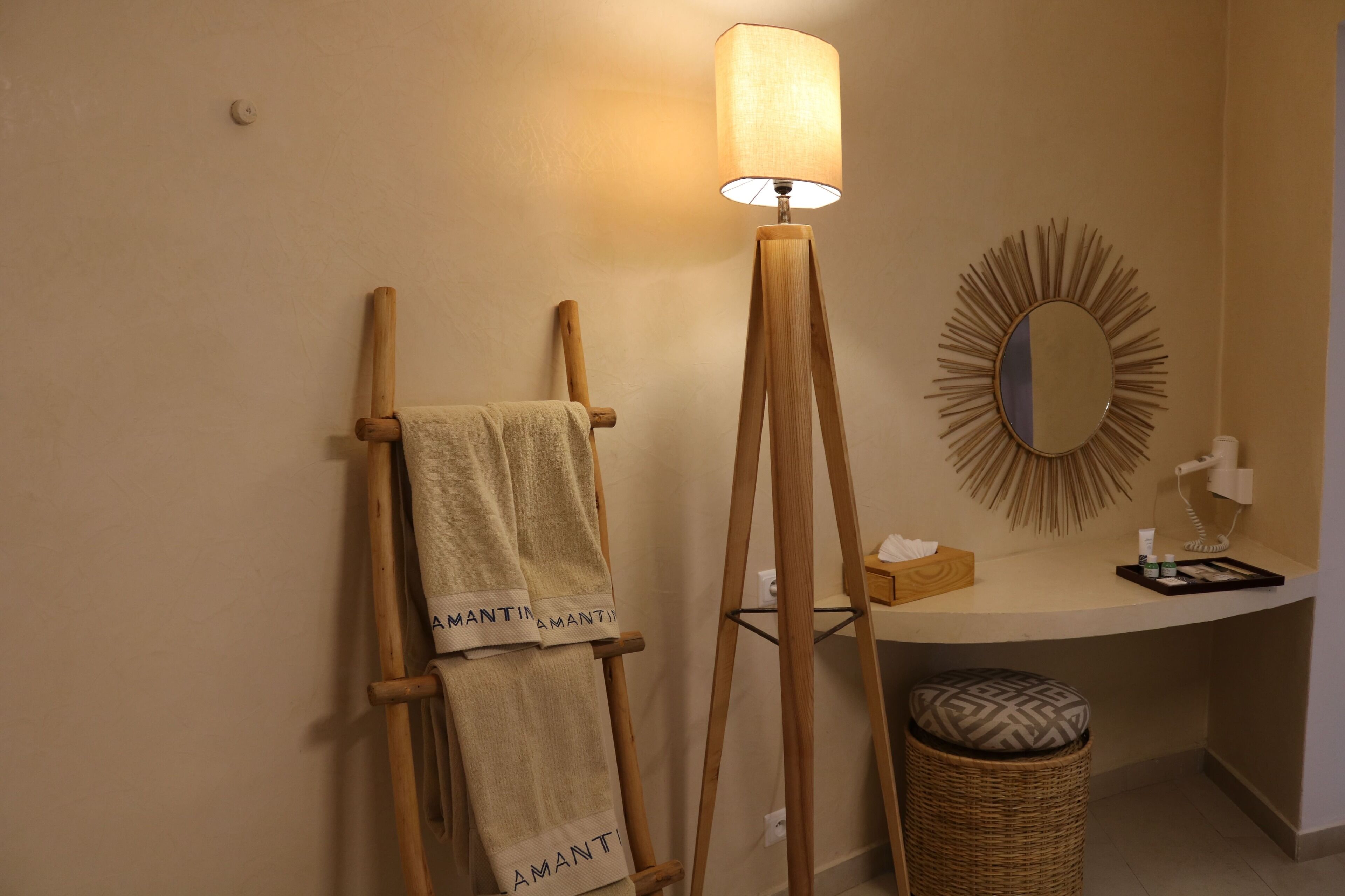 Un baño con un perchero de madera, una lámpara de pie, un espejo redondo, un taburete de mimbre pequeño y un mostrador con artículos de tocador.