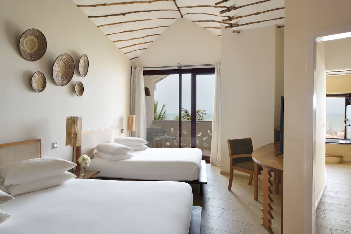 Quarto de hotel com duas camas, uma mesa de madeira, uma cadeira e varanda com vista.