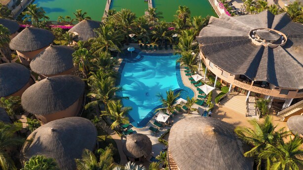 Vista aérea de un resort con una piscina central rodeada de palmeras y estructuras de techo de paja.