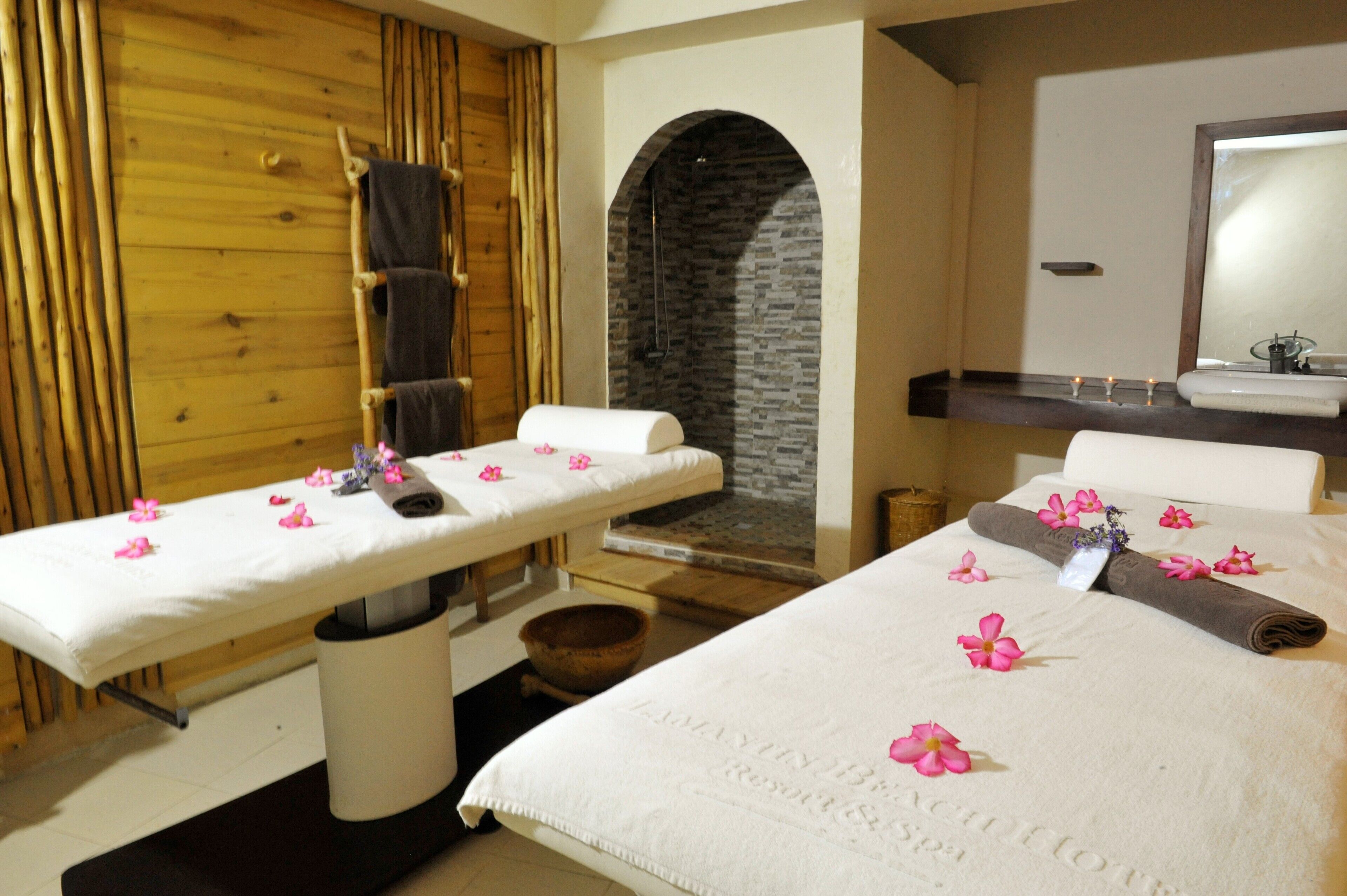 A spa room with two massage tables, a stone wall, a wooden wall, a mirror, and a sink.