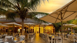 Un área de comedor al aire libre con mesas, sillas, sombrillas y palmeras.