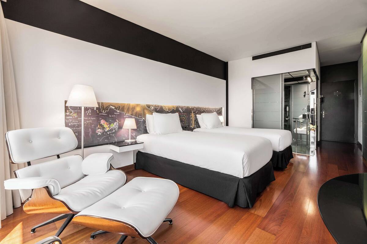 Een moderne hotelkamer met een groot bed, twee witte stoelen, een bureau en een stadsgezicht op de muur.