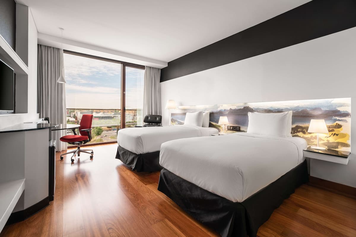 Een moderne hotelkamer met twee bedden, een bureau, een rode stoel en een groot raam met uitzicht op de stad.