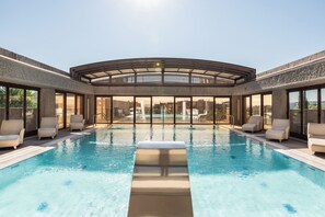 Una piscina interior moderna con paredes de vidrio transparente, varias sillas de descanso y vistas al paisaje urbano.