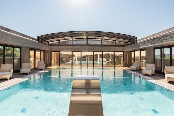 A modern indoor swimming pool with clear glass walls, multiple lounge chairs, and a view of the cityscape. - Hilton Madrid Airport (Madrid)