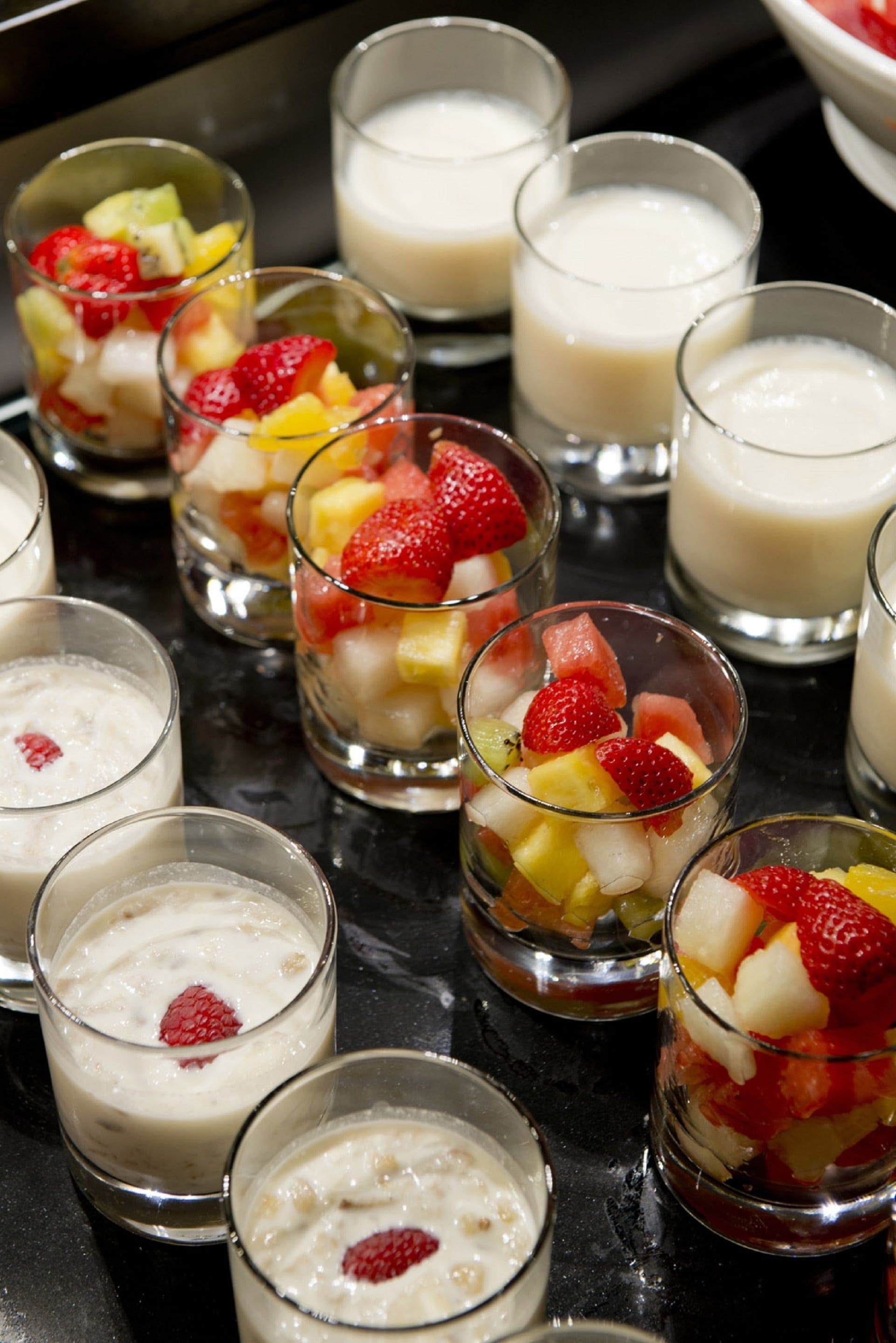 A variety of small glass dessert cups with fruit and a creamy substance, possibly yogurt or pudding, arranged on a black surface.
