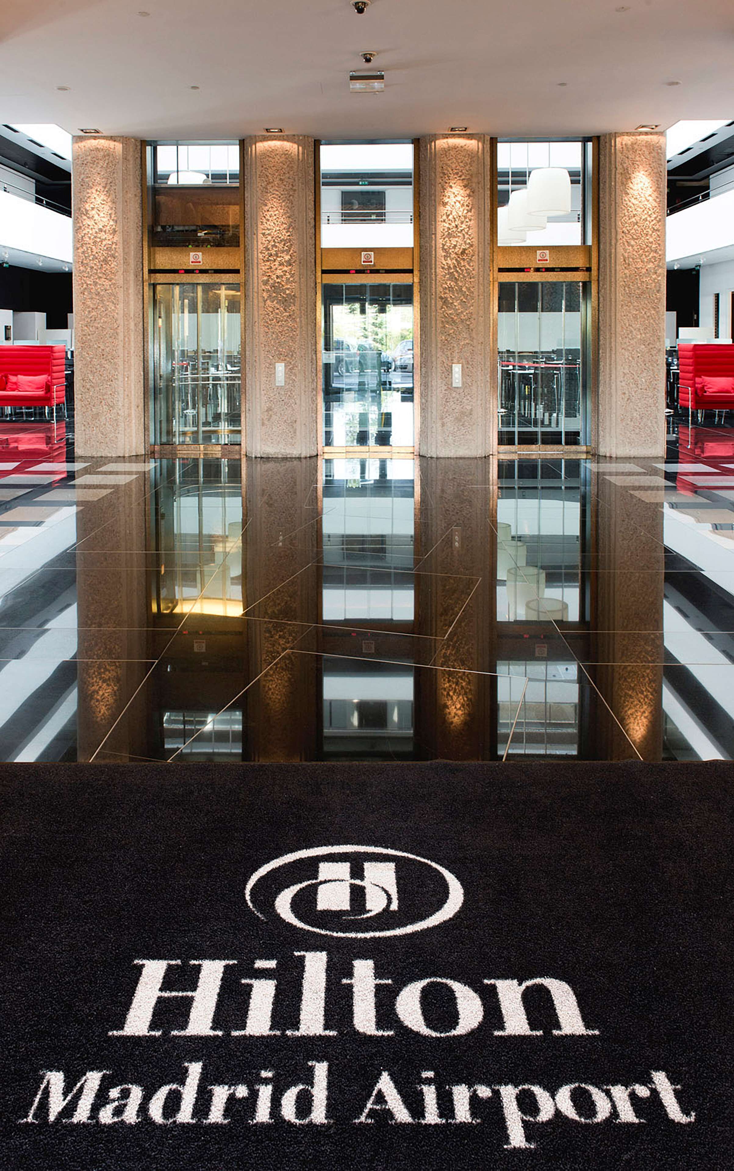 L'ingresso dell'hotel Hilton Madrid Airport, con un pavimento riflettente, sedute rosse e vista diretta sull'aeroporto sullo sfondo.