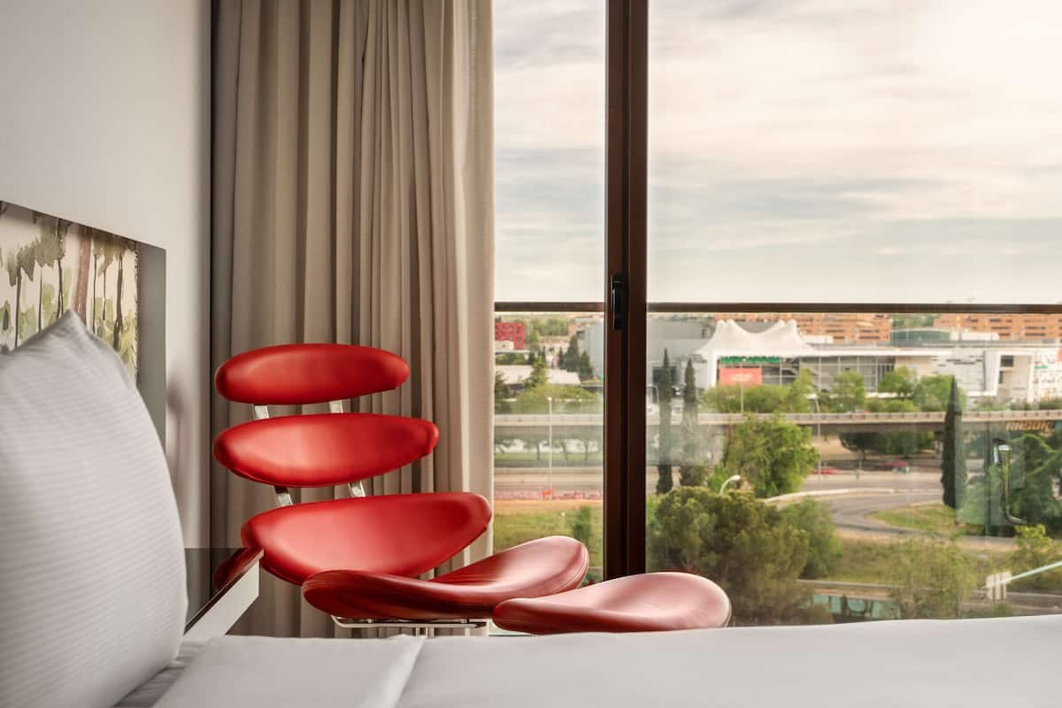 Een hotelkamer met een bed, een rode stoel, een groot raam met uitzicht op de stad en een gordijn.
