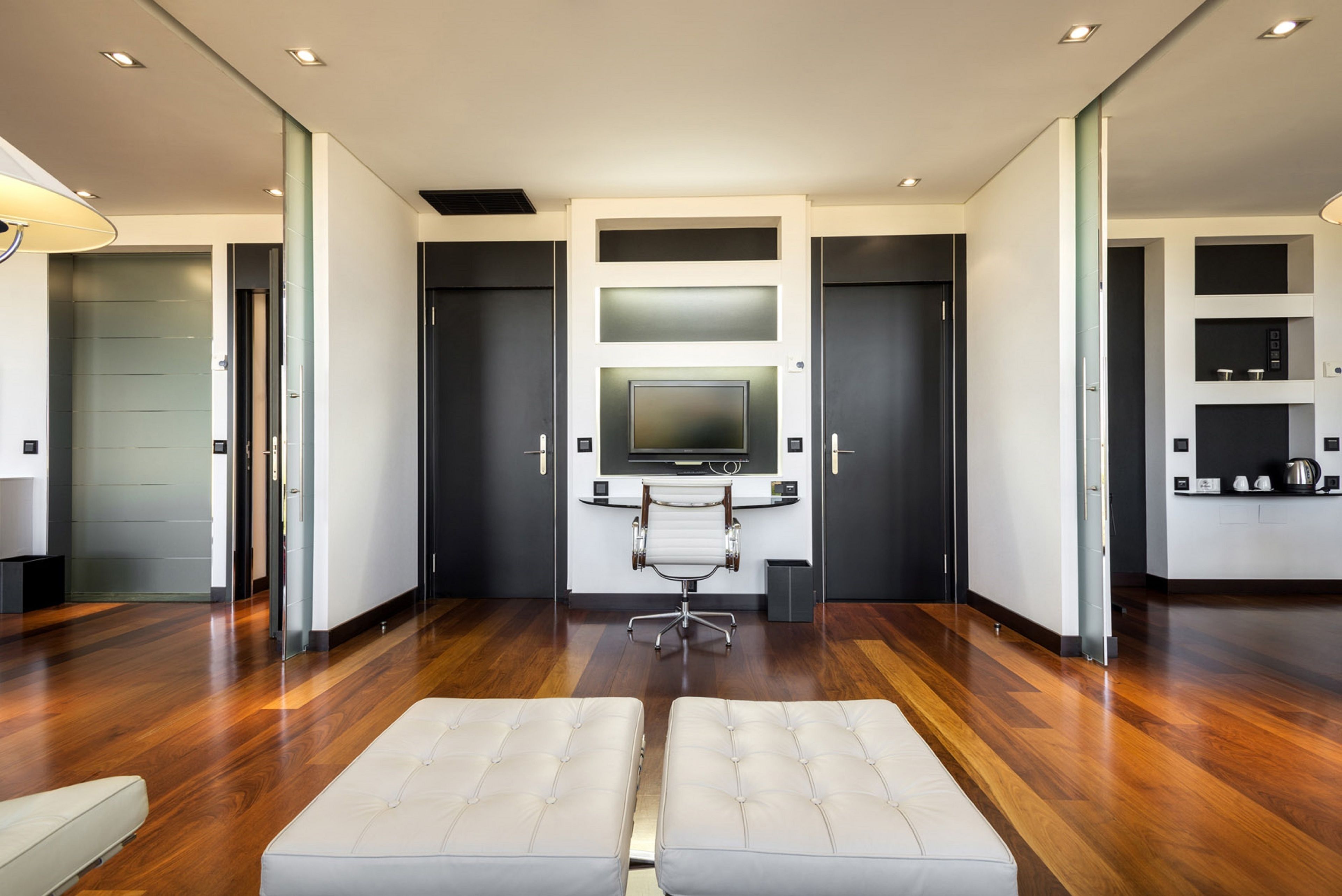 A modern hotel room with a wooden floor, a large flat-screen TV mounted on a wall, a white sofa, and a small side table.