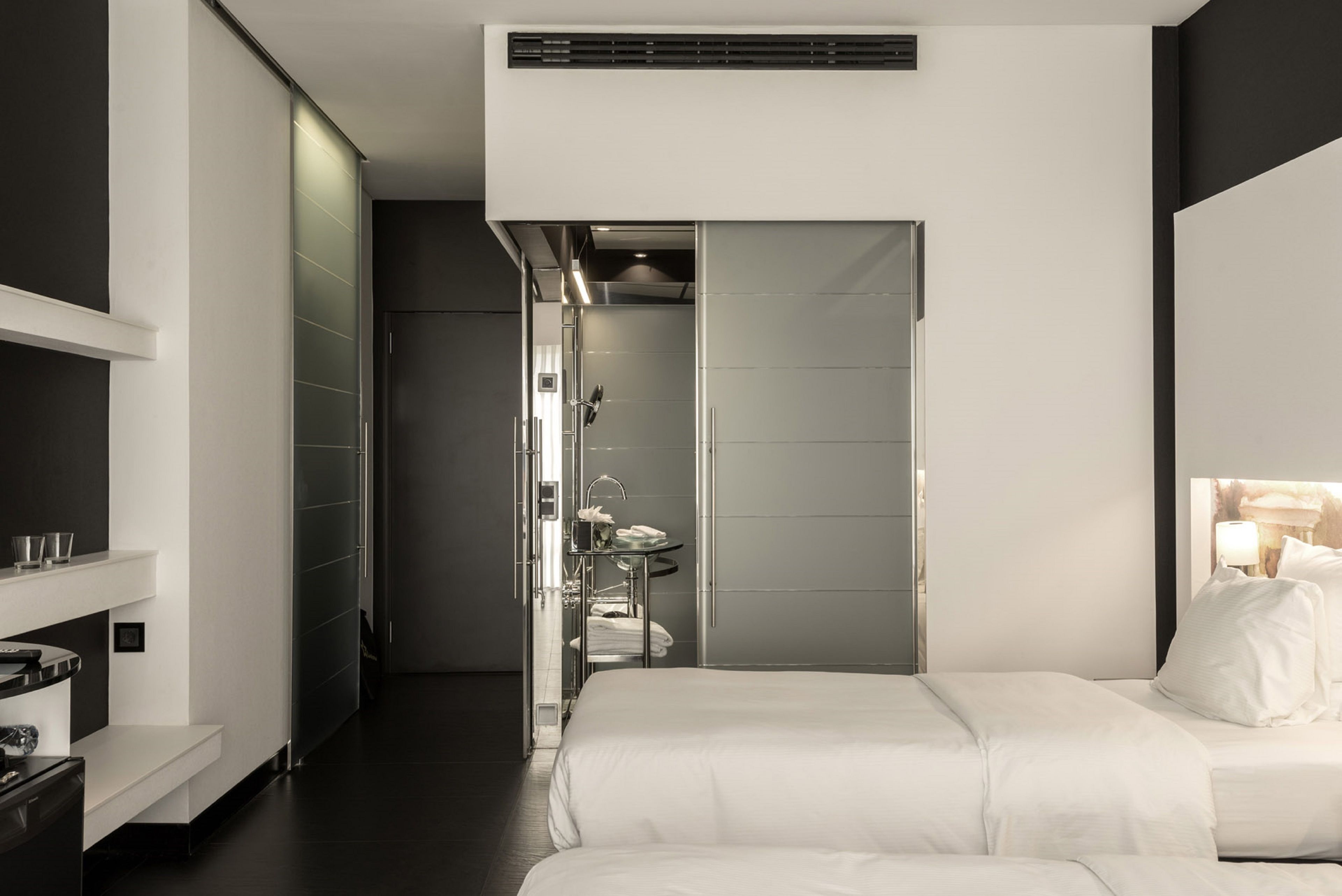 A modern hotel room with a large bed, a bedside table, a built-in wardrobe, and a bathroom with a shower and sink.