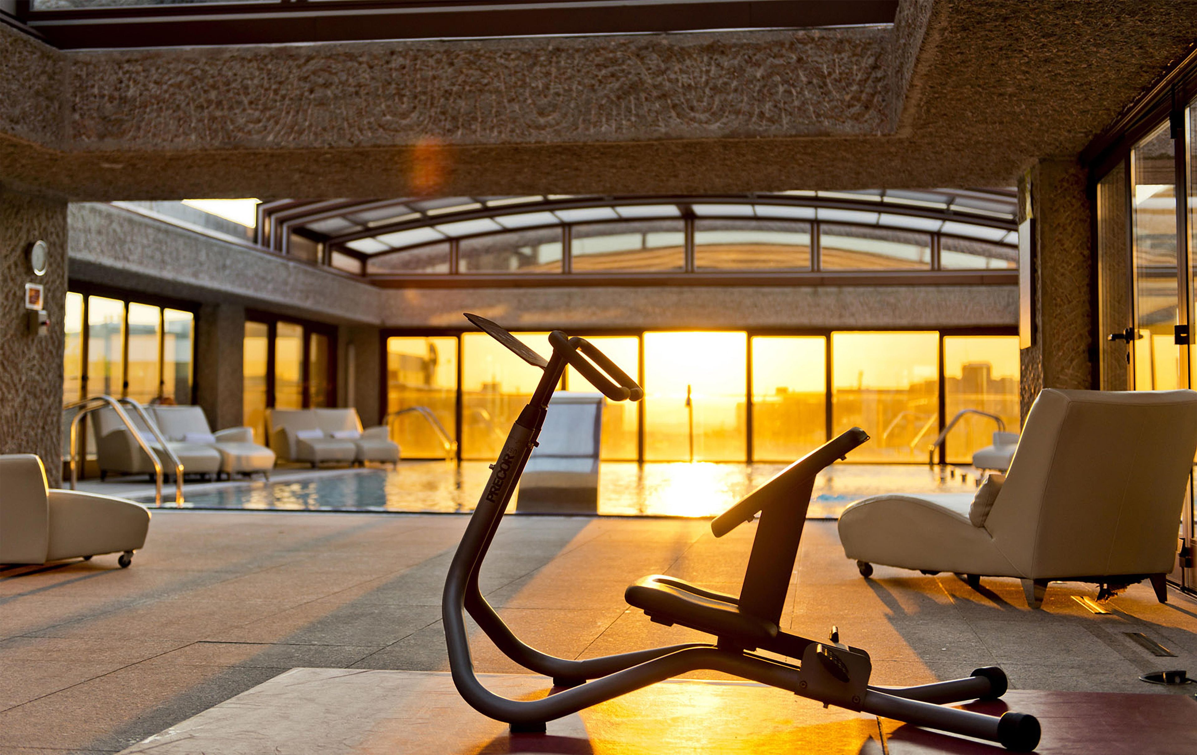 Un espacio interior de ejercicio con una bicicleta estática, sillones de descanso y una piscina al fondo.