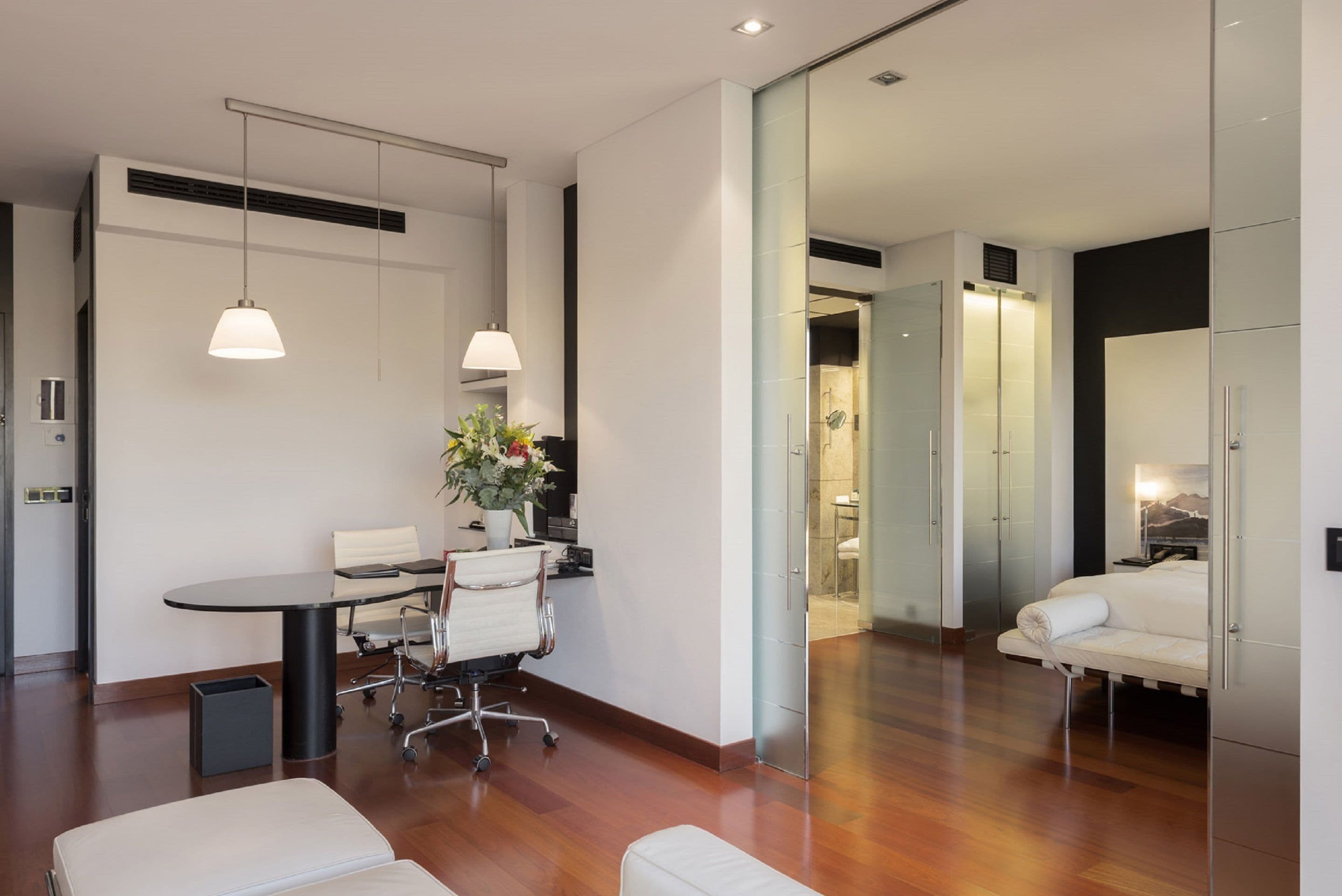 Una habitación de hotel moderna con suelo de madera, un área de comedor con mesa y sillas, un sofá y un espejo grande.