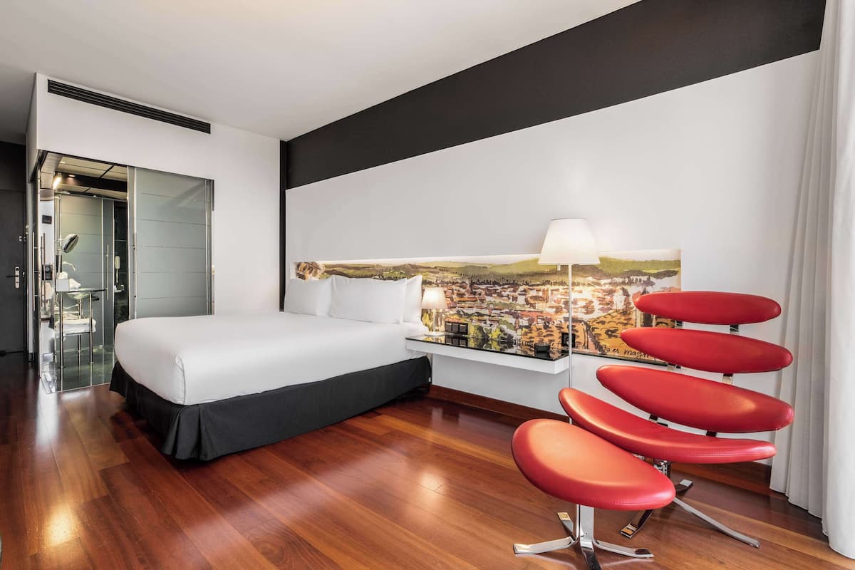 Een moderne hotelkamer met een groot bed, een badkamer met glazen scheiding, een stadslandschap schilderij en een rode stoel.