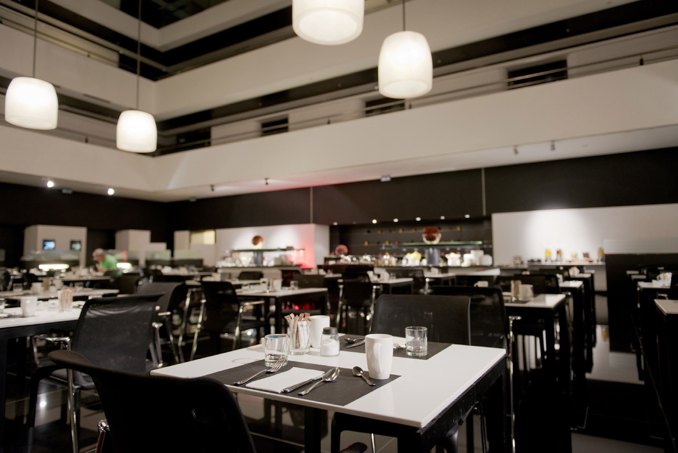 Um restaurante moderno com várias mesas e cadeiras, uma área de cozinha ao fundo e iluminação pendente.