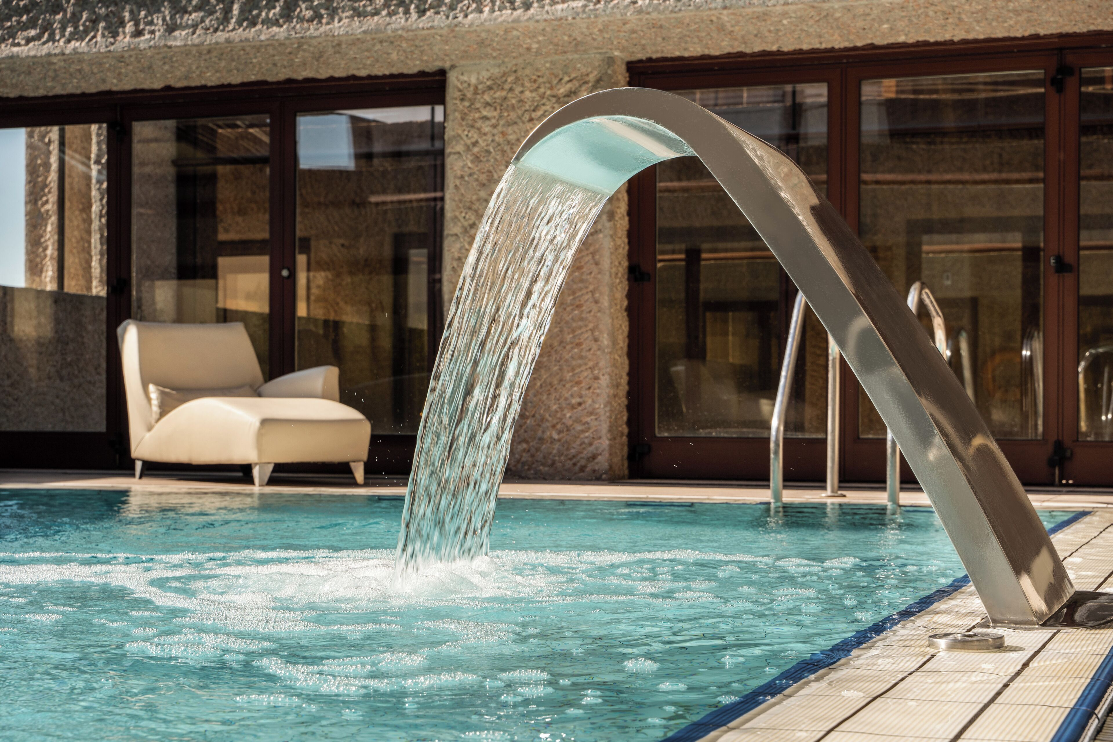 Una piscina con fuente, una silla de descanso y un edificio con ventanales grandes.
