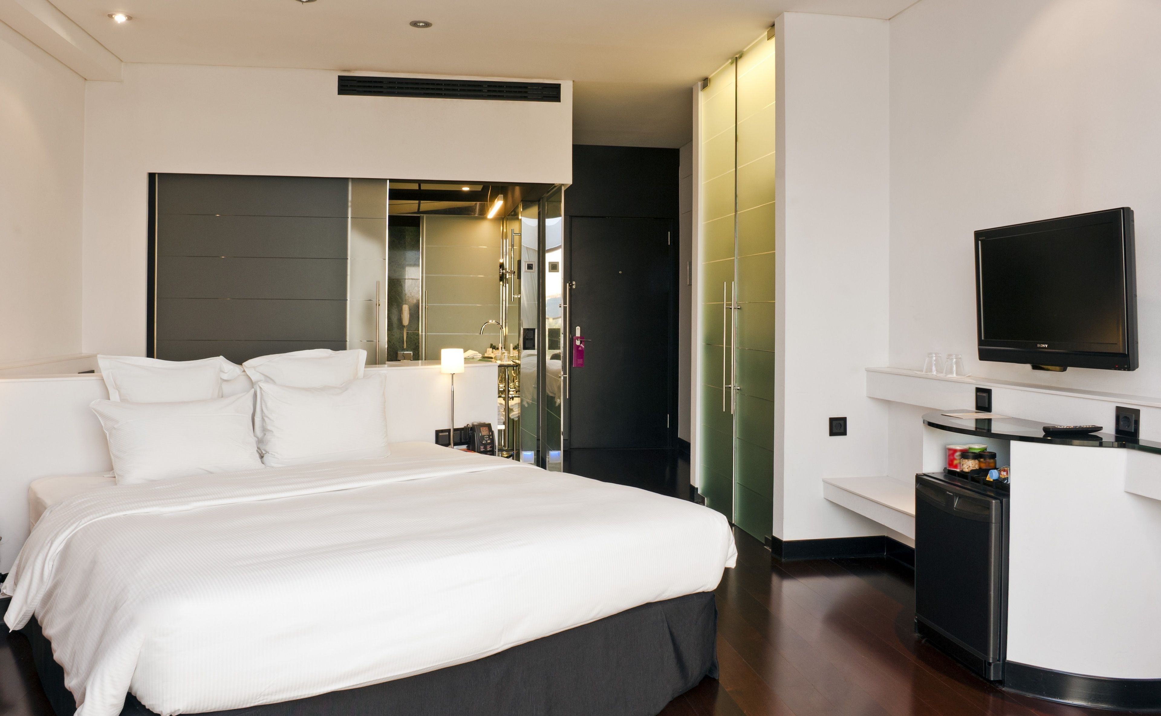 A hotel room with a bed, a TV, a minibar, and a bathroom with a mirror and sink.