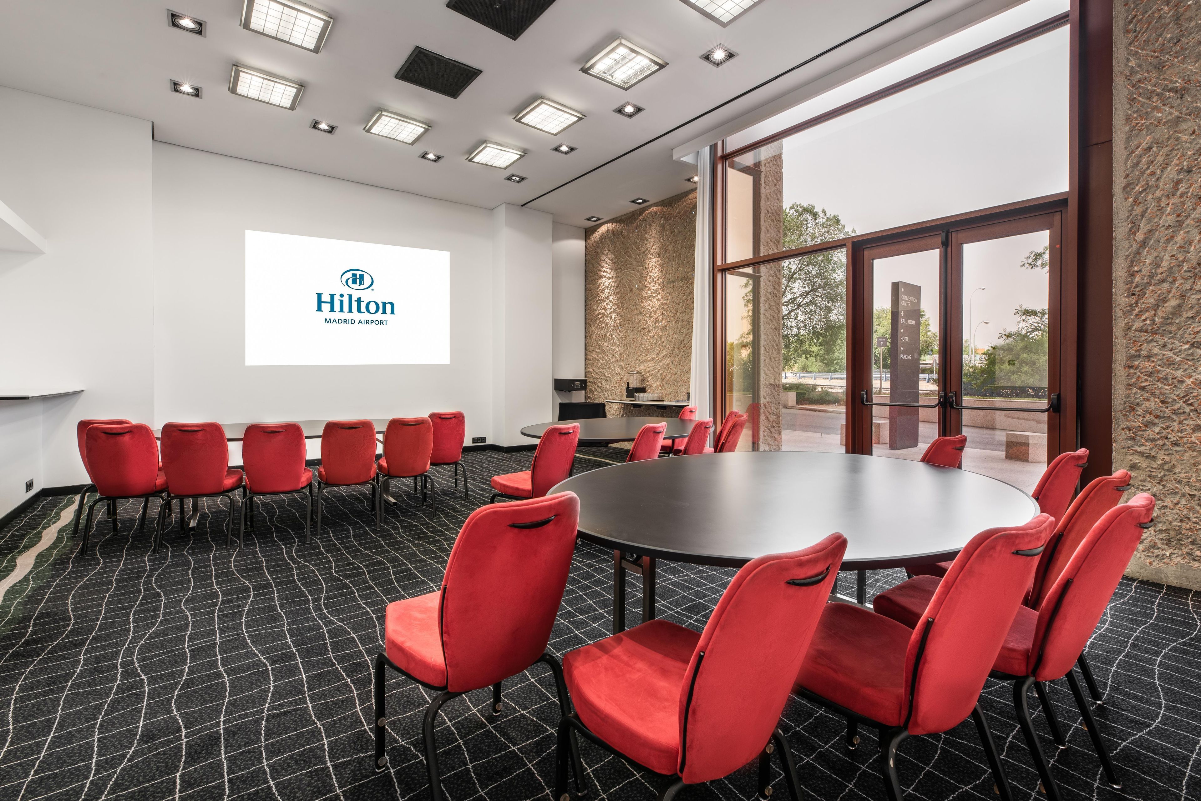 A conference room with a large table and red chairs, a projector screen with the Hilton logo, and a view through the glass door.
