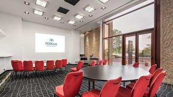 A conference room with a large table and red chairs, a projector screen with the Hilton logo, and a view through the glass door.