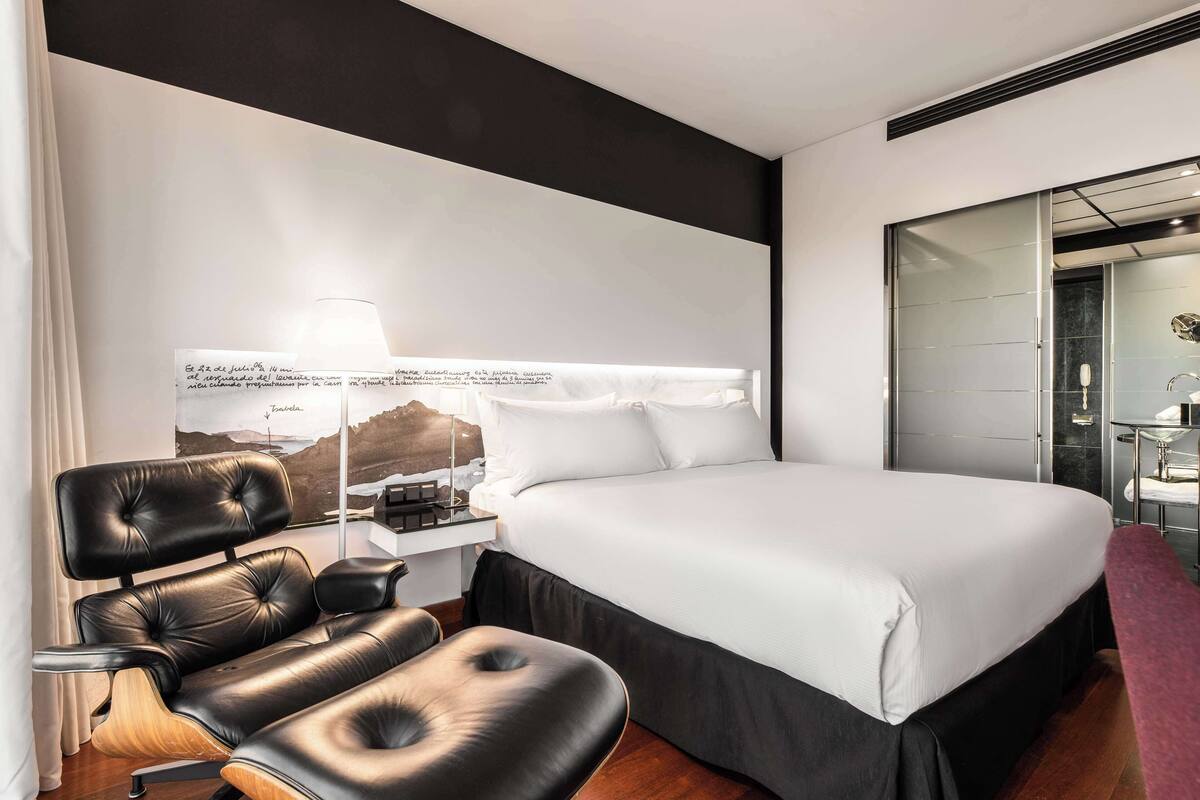 Een moderne hotelkamer met een groot bed, een zwarte leren fauteuil, een nachtkastje met lamp en uitzicht op de bergen door het raam.