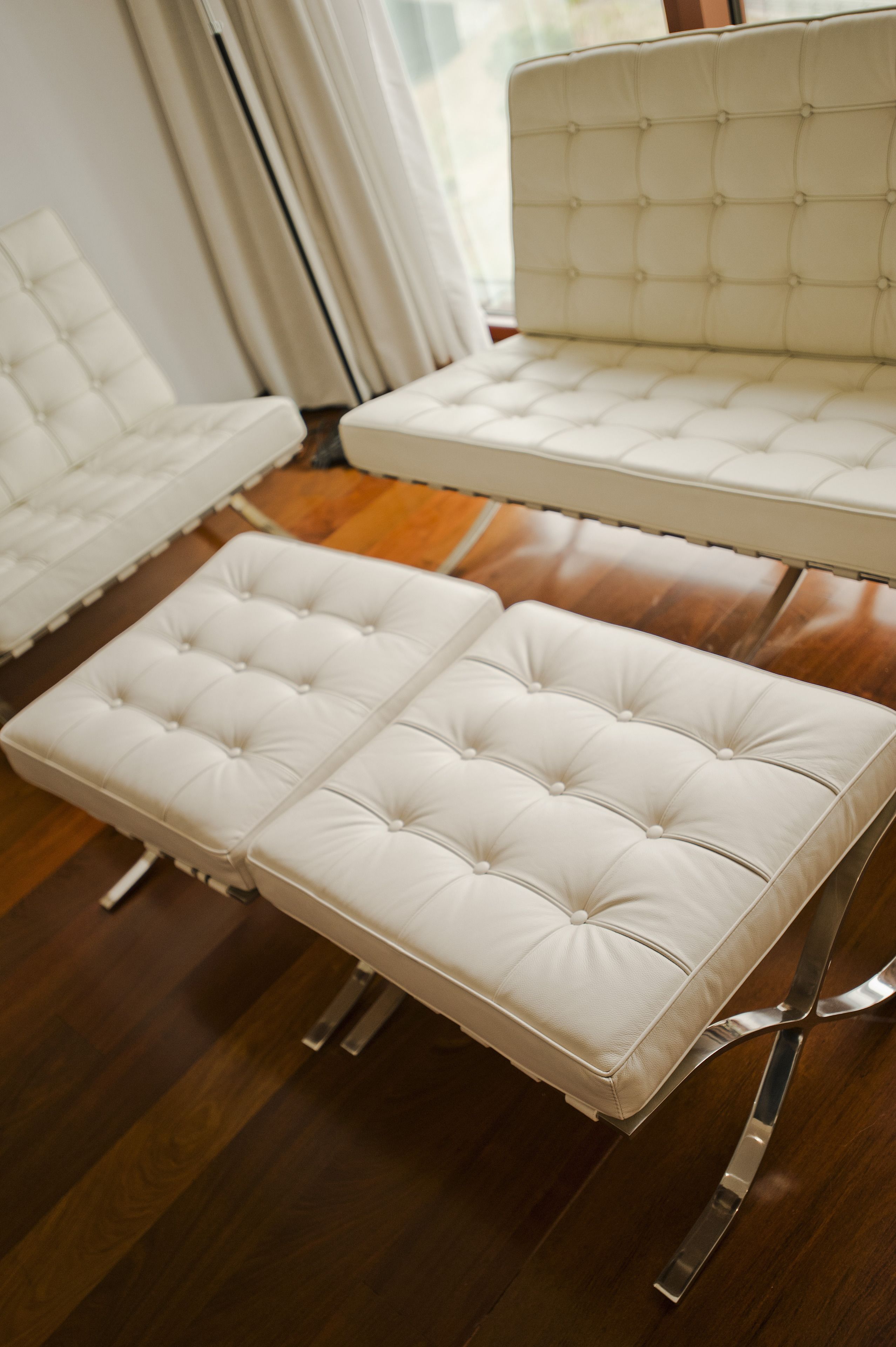 A modern living room with a white tufted sofa and ottoman, wooden flooring, and a large window with curtains.