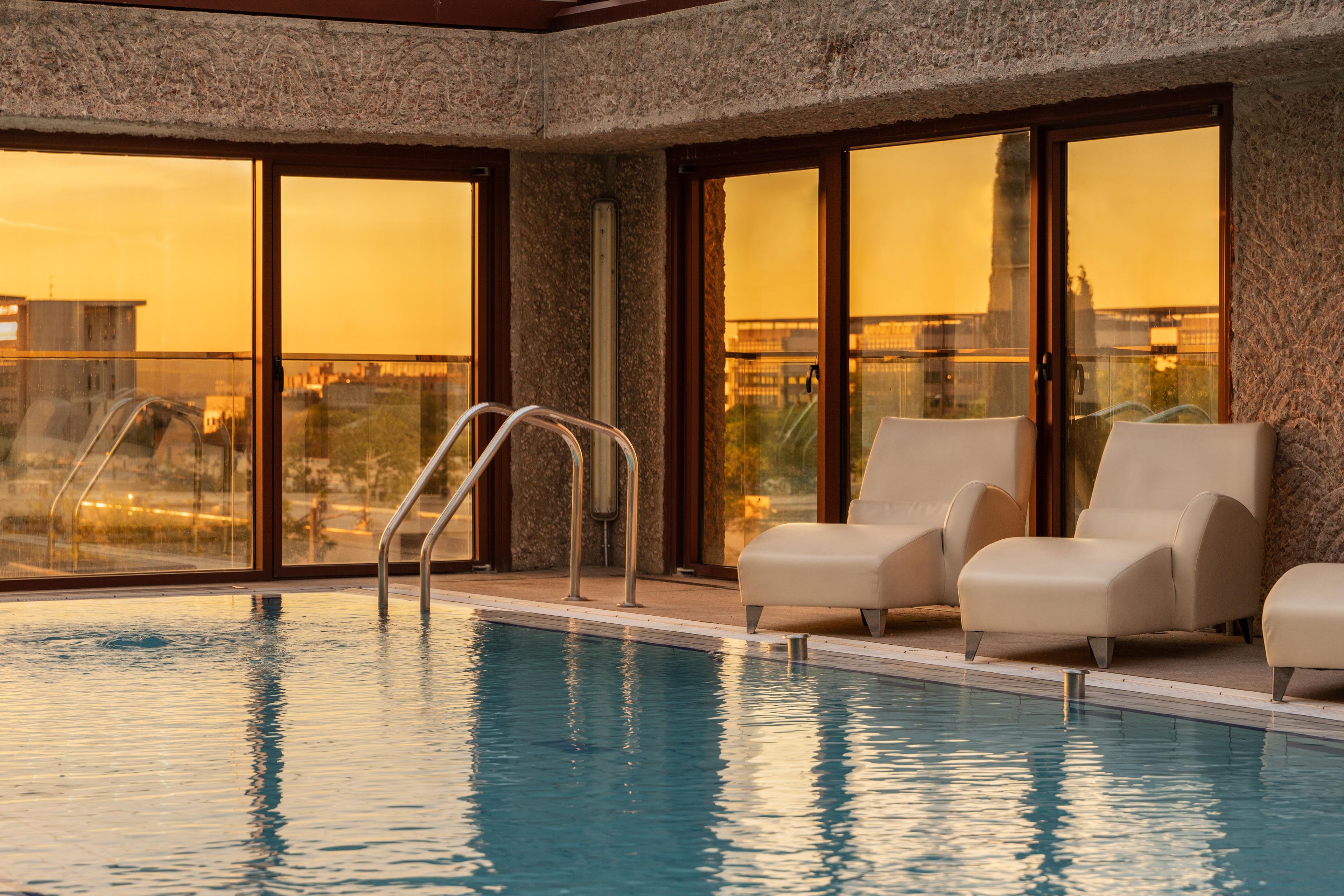 An indoor swimming pool with clear glass walls, a metal ladder, and two lounge chairs.