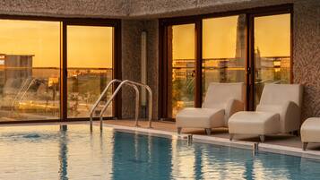An indoor swimming pool with clear glass walls, a metal ladder, and two lounge chairs.