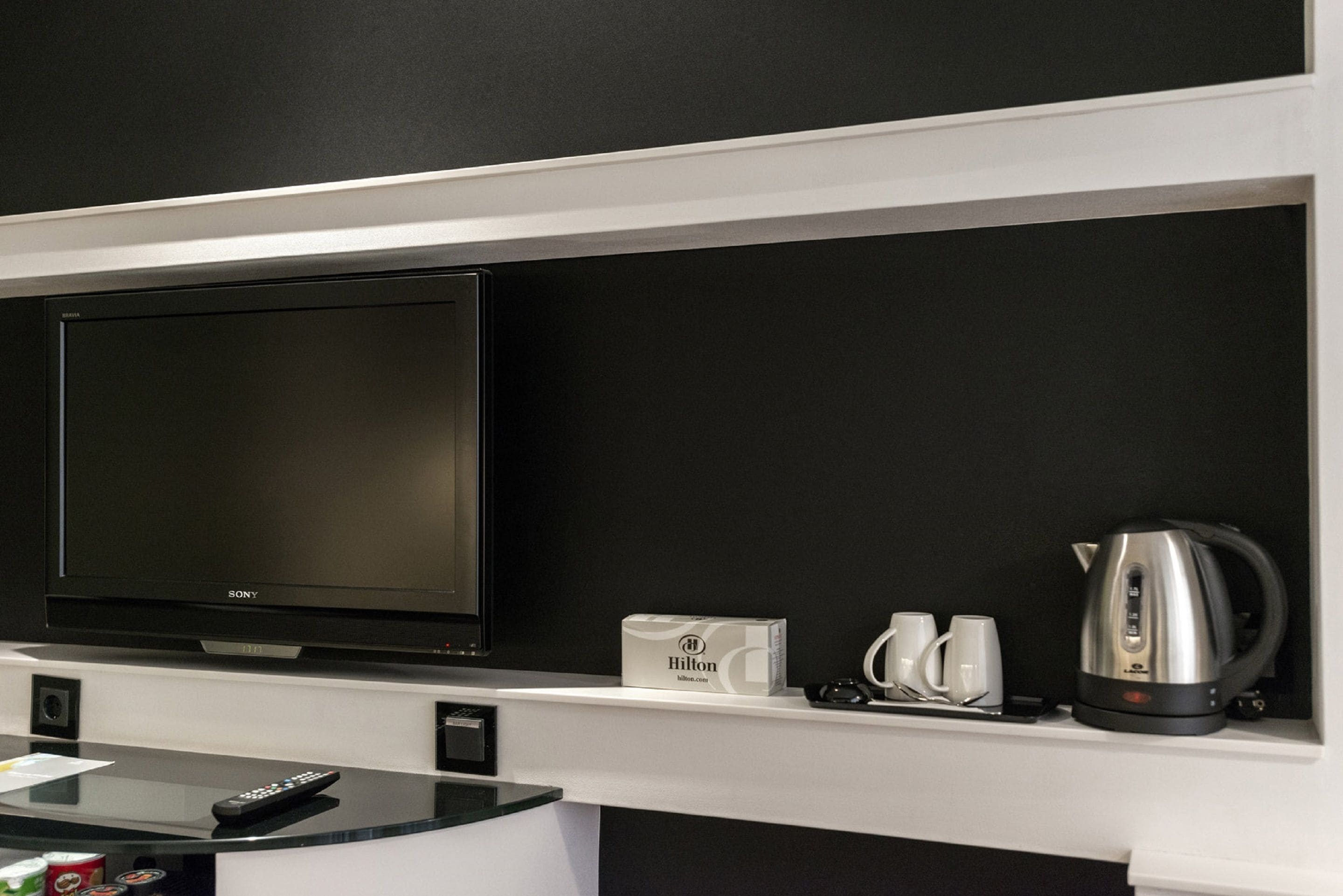 Habitación de hotel con televisión de pantalla plana, minibar, hervidor y juego de tazas blancas.
