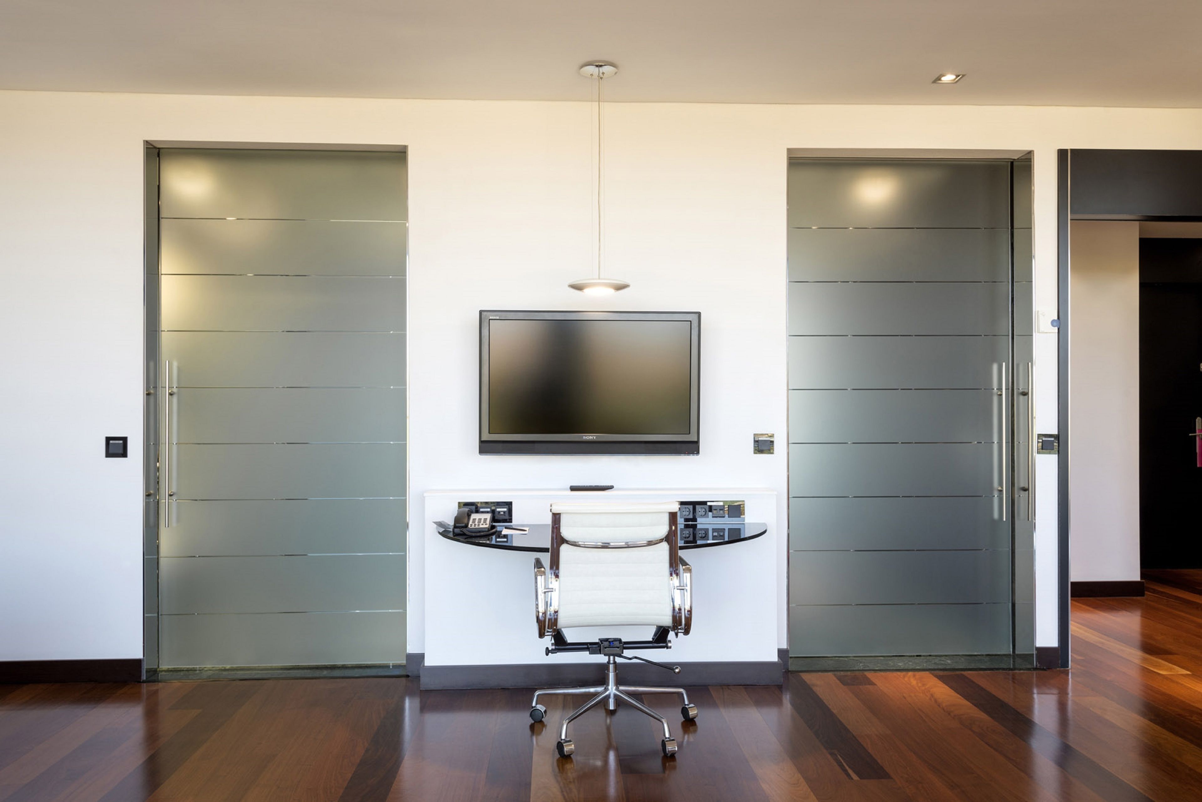 A modern office space with a large flat-screen TV mounted on the wall, a white office chair, and a desk with a computer.