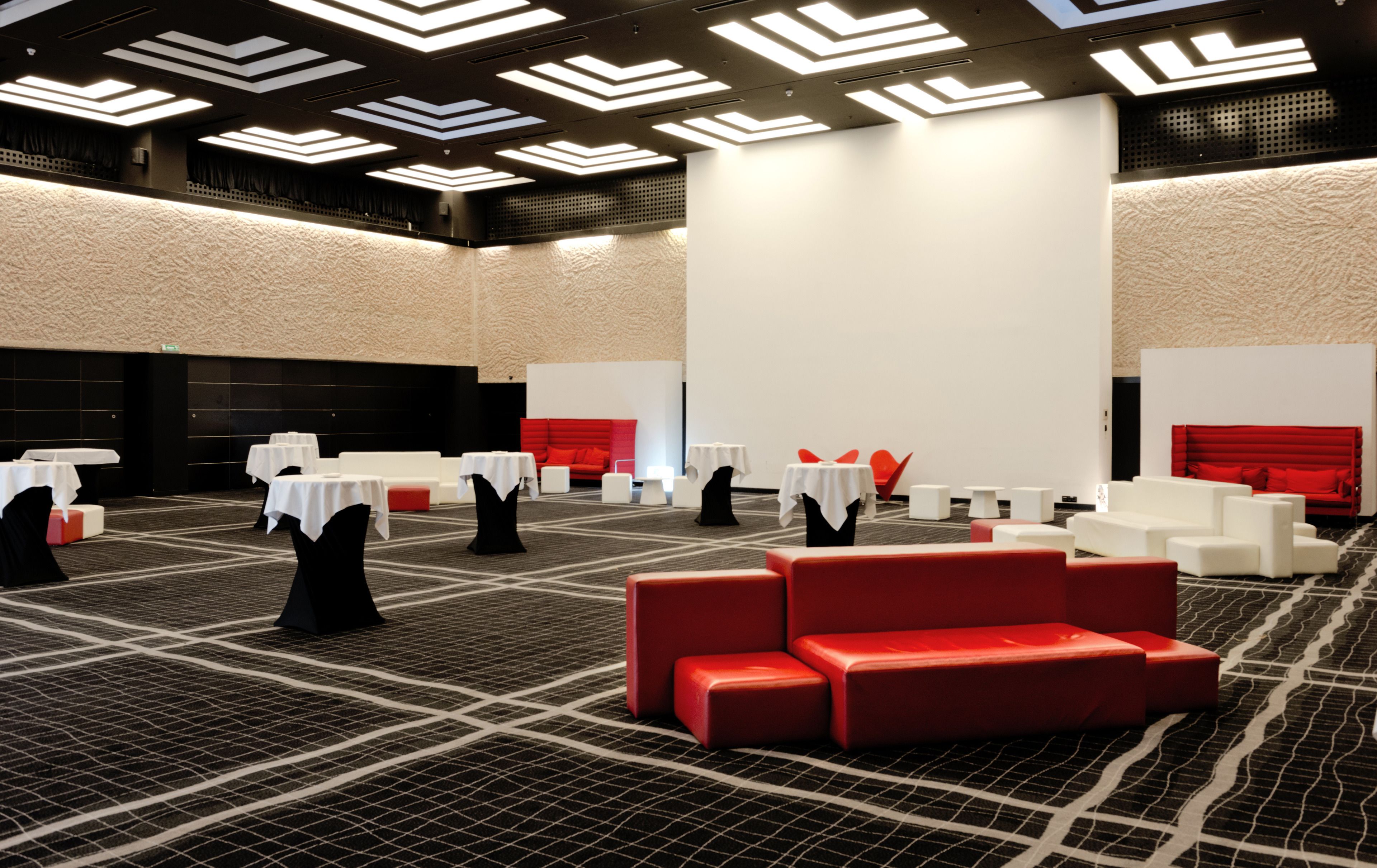 Een moderne hotellobby met een geometrisch patroon in het tapijt, witte tafels met zwarte stoelen, rode banken en een hoog plafond met lineaire verlichting.
