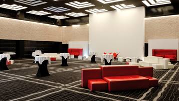 Een moderne hotellobby met een geometrisch patroon in het tapijt, witte tafels met zwarte stoelen, rode banken en een hoog plafond met lineaire verlichting.
