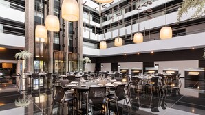 Un espace de restauration spacieux et moderne, doté de nombreuses tables et chaises, de grands luminaires suspendus et d’un sol noir poli.