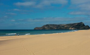 A wide sandy beach with footprints leading towards the water, a rocky outcrop in the distance, and a clear blue sky. - Torre Praia Hotel (Porto Santo)