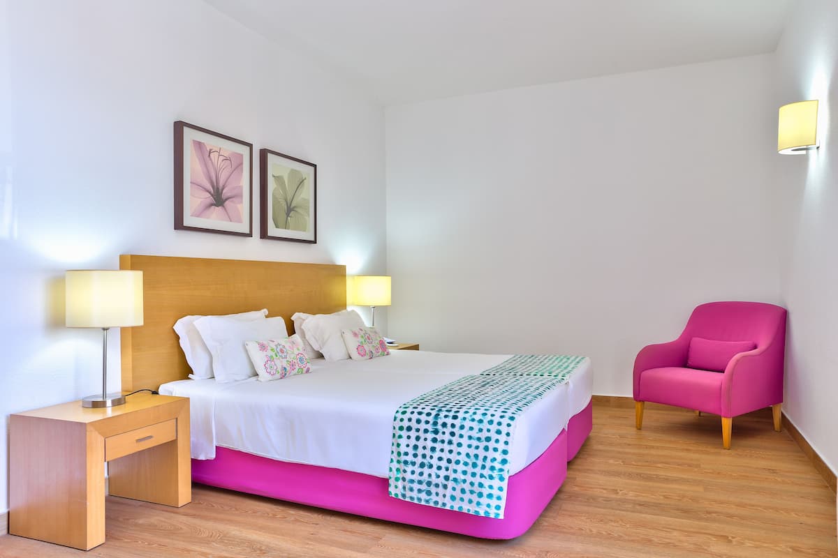 Habitación de hotel con cama, mesita de noche, lámpara, silla y dos cuadros colgados en la pared.