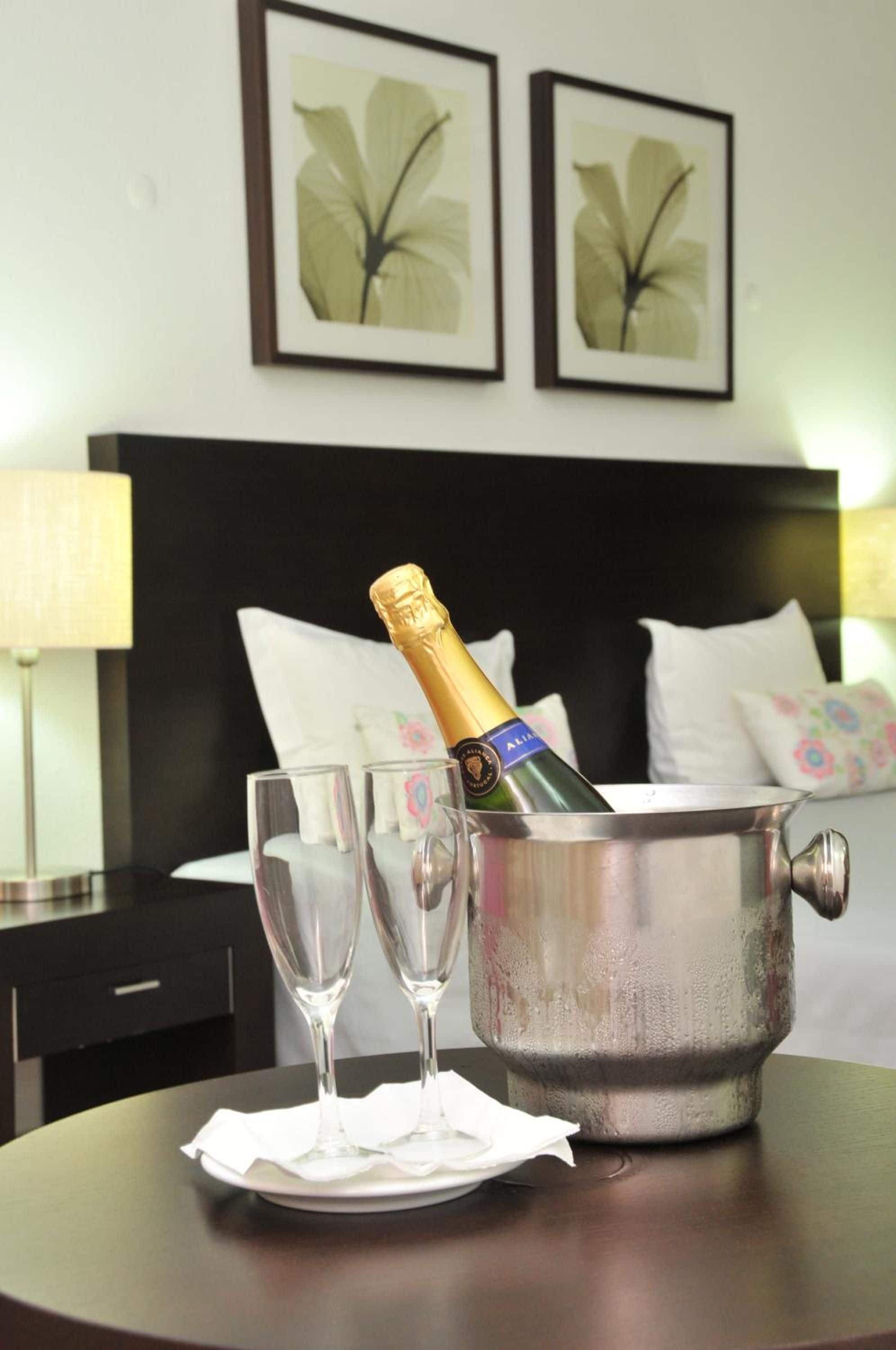Una habitación de hotel con una cama, una mesita de noche, una lámpara, dos cuadros enmarcados con motivos florales en la pared, una mesa con una botella de champán y dos copas.