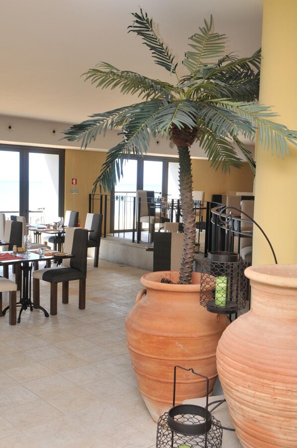 A spacious indoor area with large terracotta pots, a palm tree, and a view of the sea through large windows.