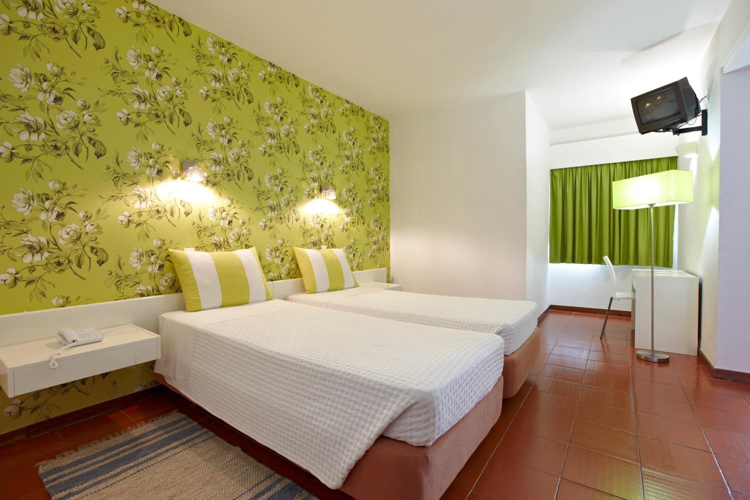 A hotel room with two beds, a green floral wallpaper, a white nightstand, a lamp, and a television.
