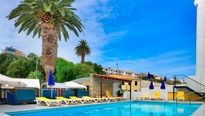 A swimming pool with sun loungers and umbrellas, surrounded by palm trees and a building with a tiled roof. - Praia Dourada Hotel (Porto Santo)