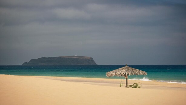 A beach with a thatched umbrella and a distant island. - Praia Dourada Hotel (Porto Santo)