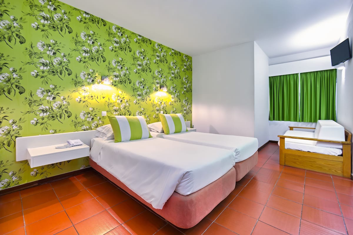 Una habitación de hotel con dos camas, una mesita de noche pequeña, una silla, un televisor y cortinas verdes.