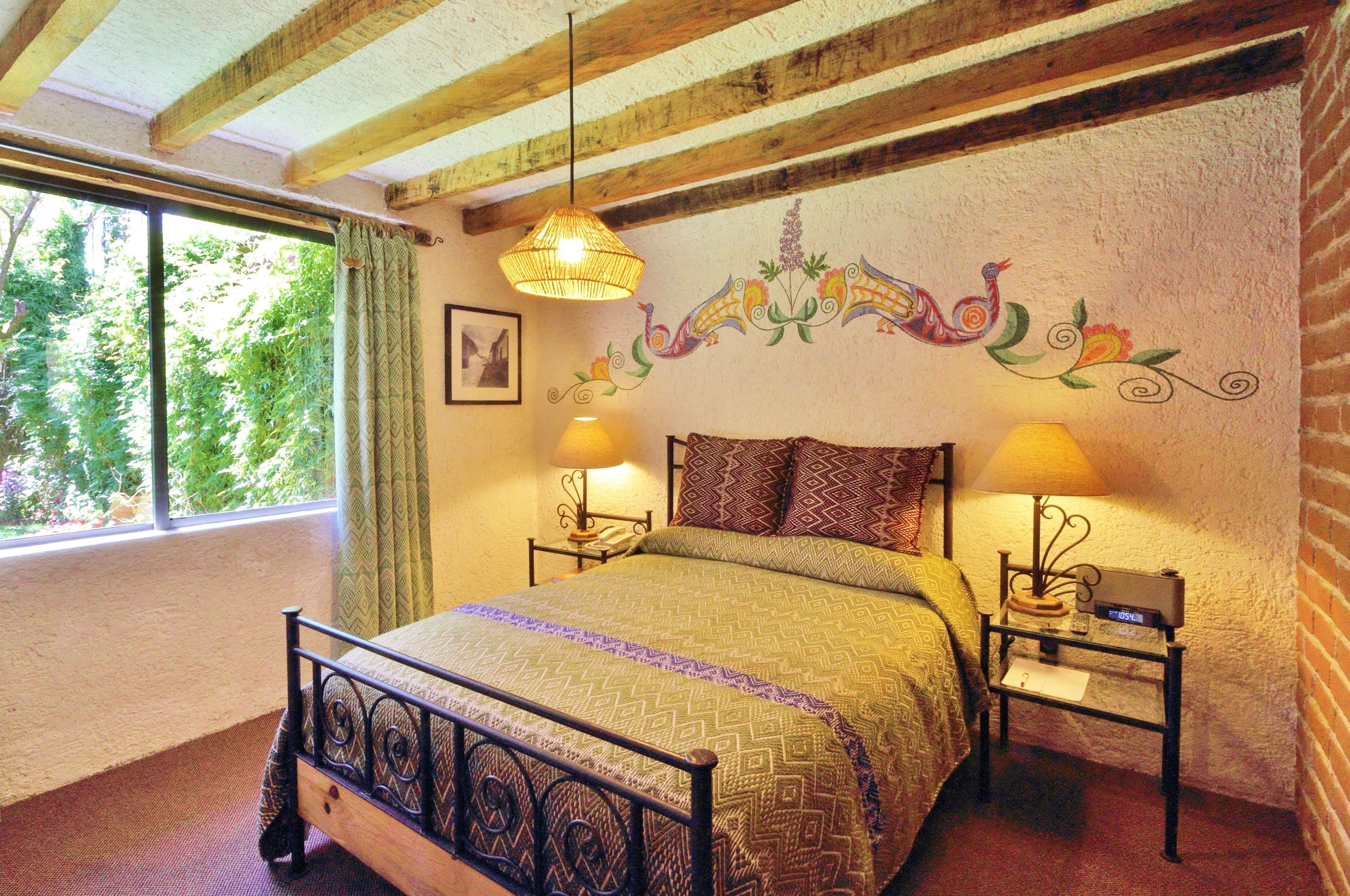 A bedroom with a bed, two bedside tables with lamps, a window with curtains, and a decorative wall mural.