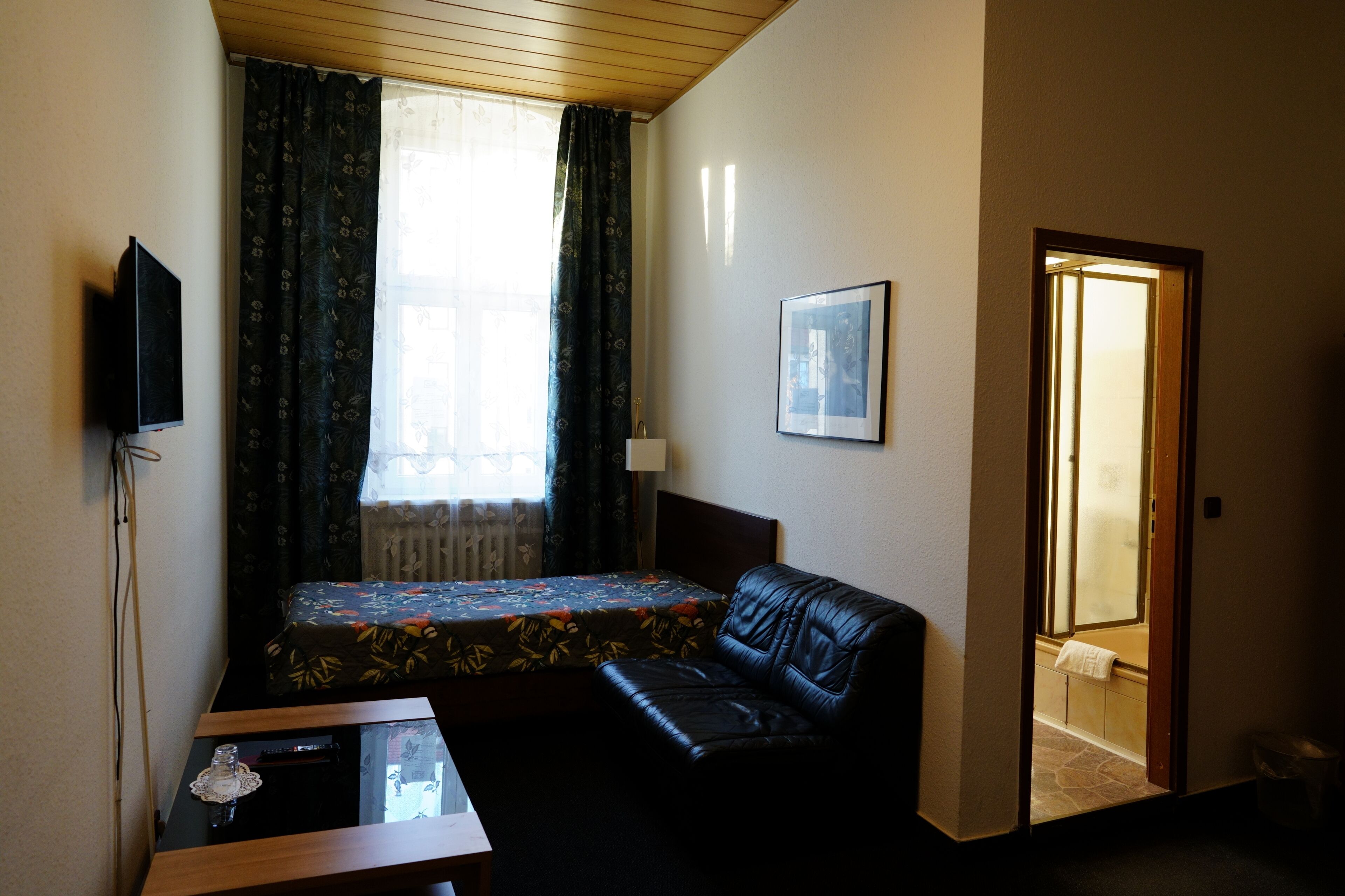Una habitación pequeña y tenuemente iluminada, con una cama individual, un sofá de cuero, un televisor y una pequeña mesa con un objeto de vidrio encima.