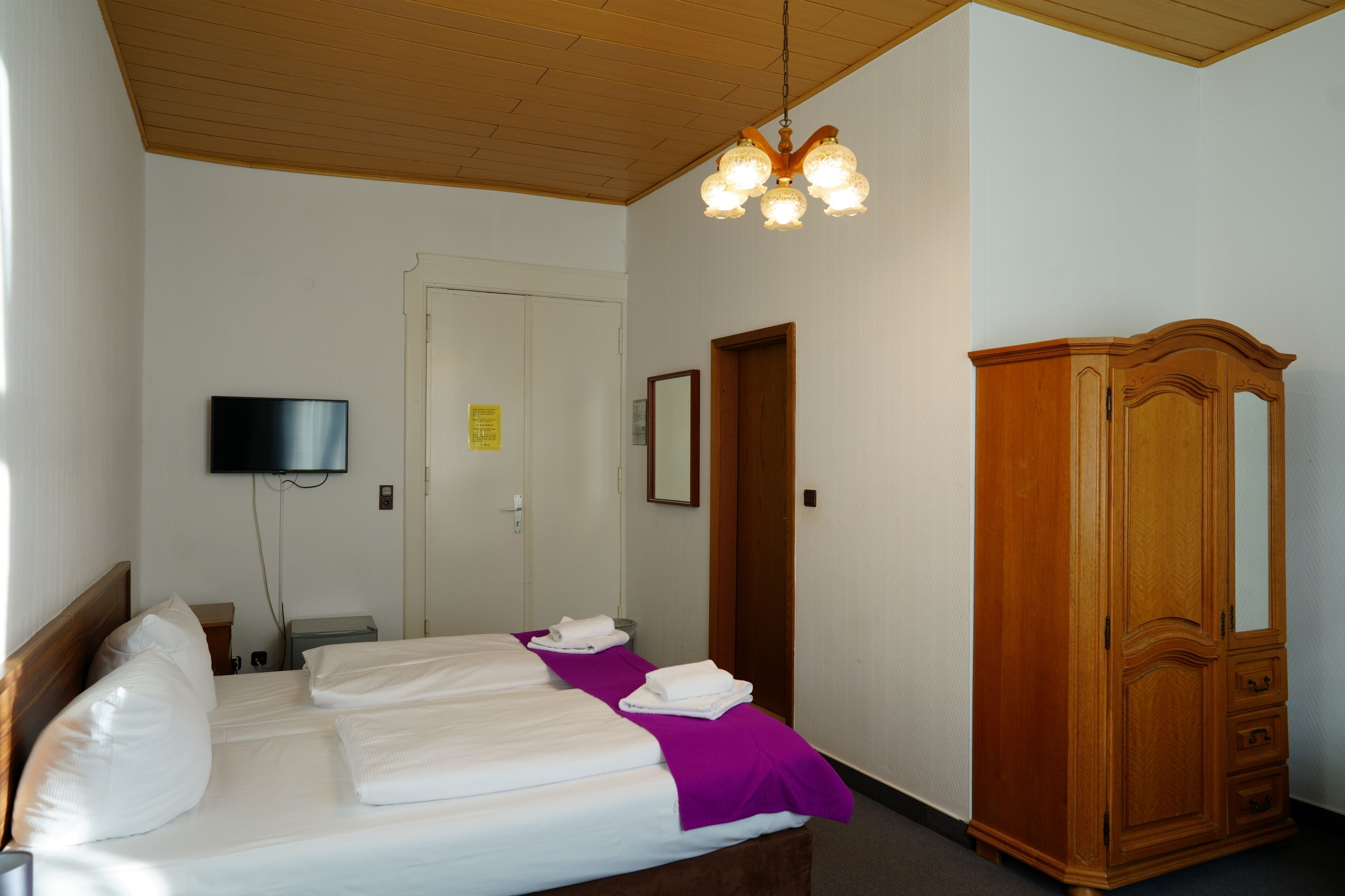 A hotel room with a bed, a wooden wardrobe, a television on the wall, and a ceiling light fixture.