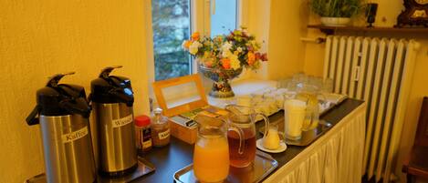 Montaje de desayuno tipo buffet con dispensadores de café y jugo, un bol de fruta y una bandeja con diversos productos para el desayuno.