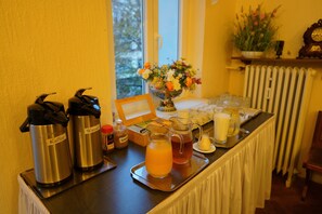 A breakfast buffet setup with coffee and juice dispensers, a fruit bowl, and a tray with various breakfast items. - Hotel Margrit (Berlin)