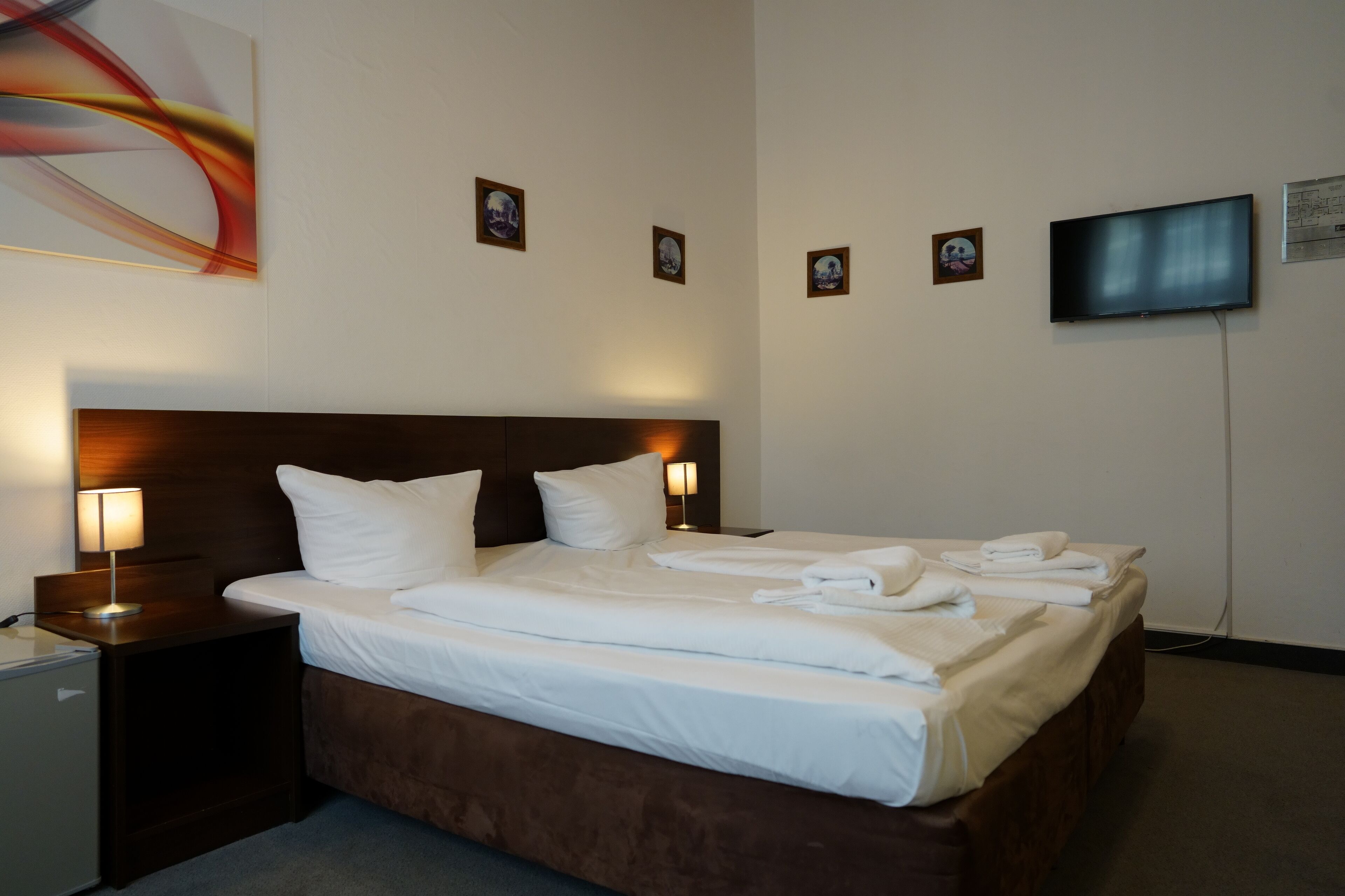 A hotel room with a large bed, a TV mounted on the wall, and a small desk with a lamp.