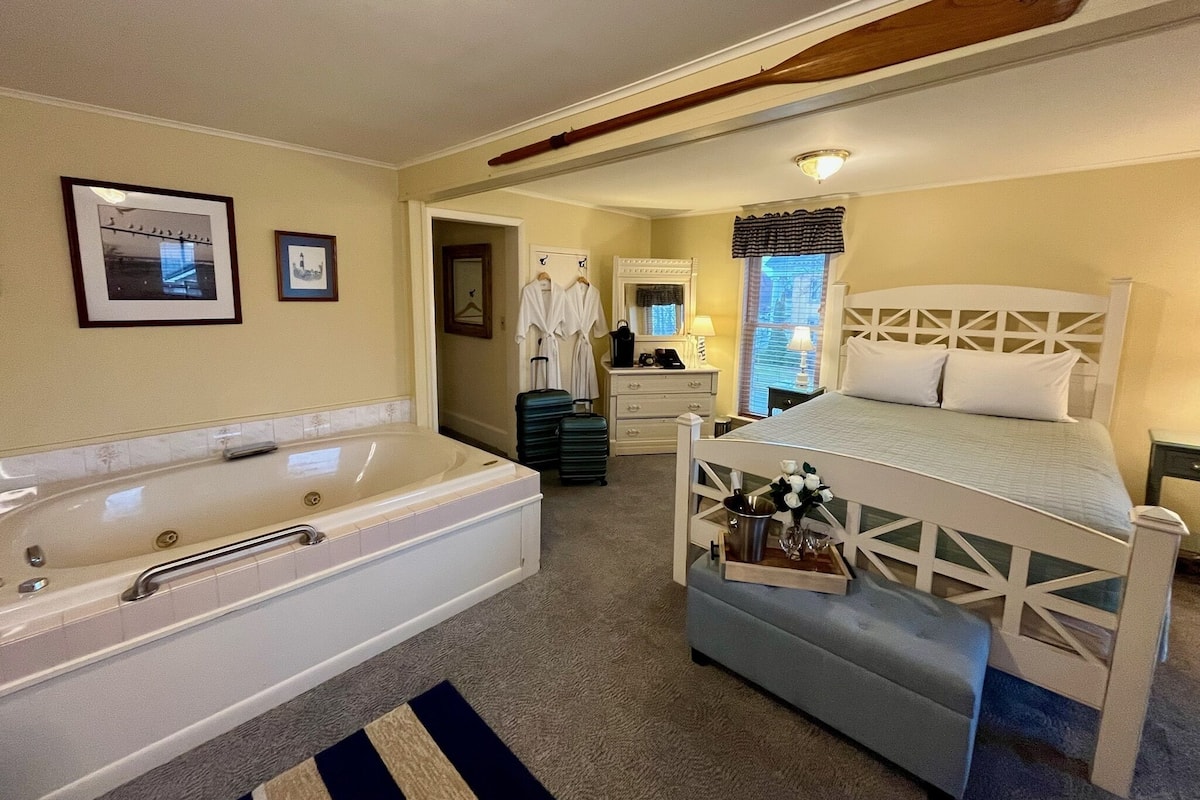 A bedroom with a large white bathtub, a bed with a white headboard, and a small bench. There are two framed pictures on the wall, a mirror, and a window with blinds.