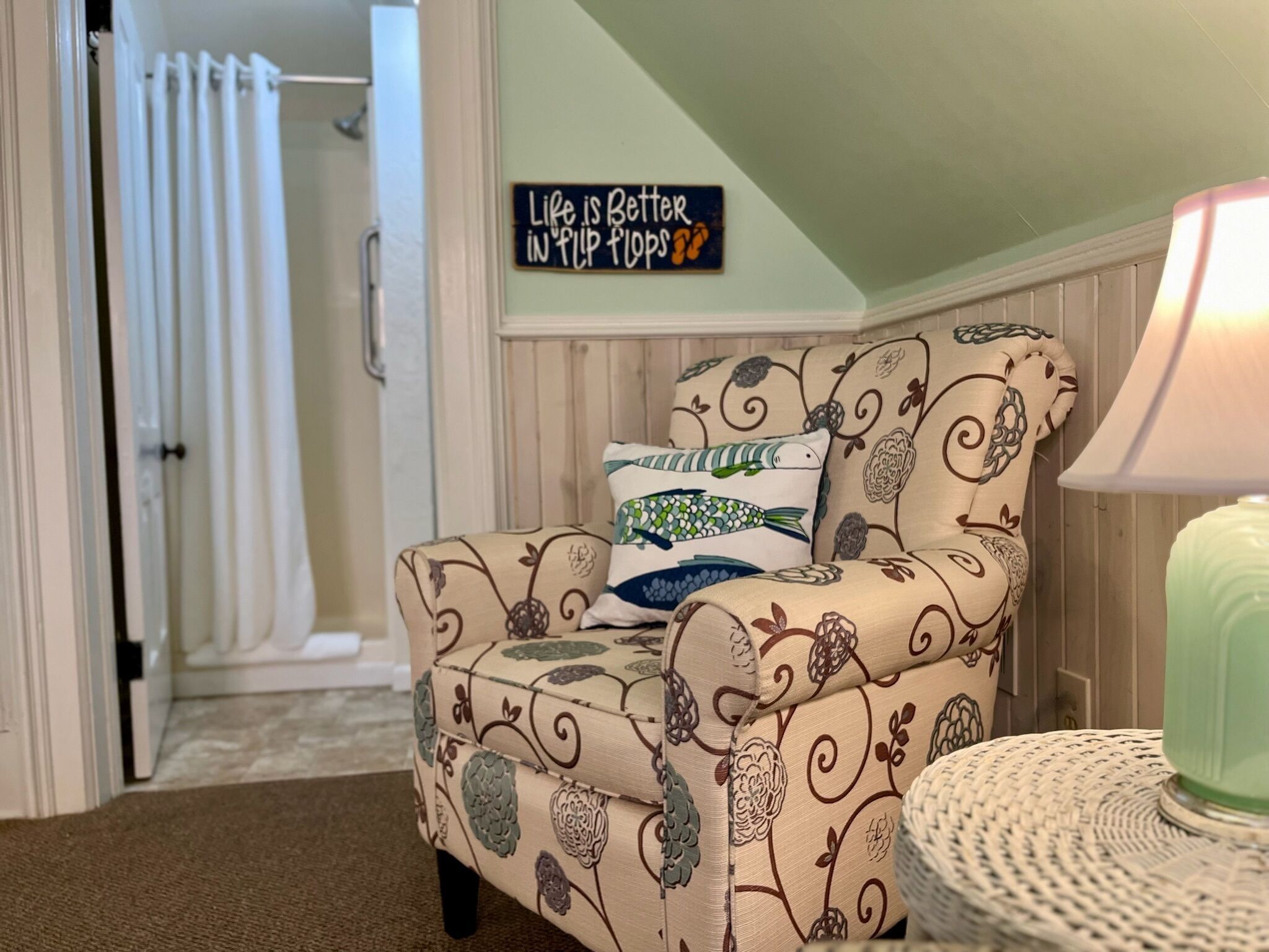 A cozy living room with a patterned armchair, a decorative pillow, a wall sign, and a lamp.